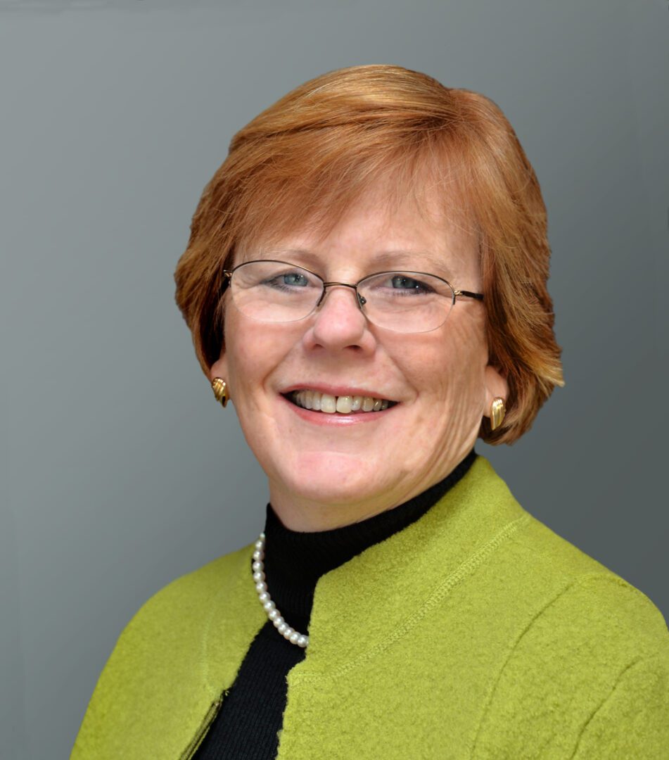 Smiling woman with glasses and short red hair wearing a green jacket.